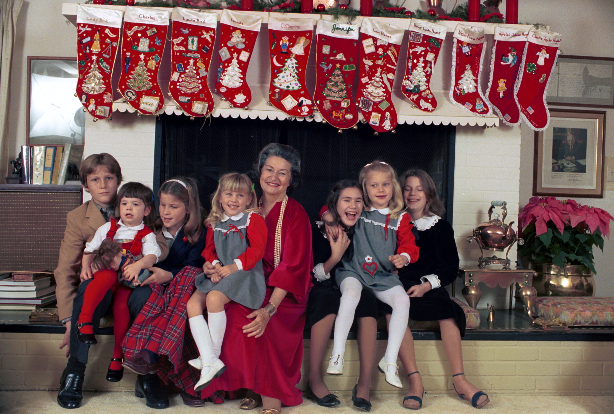 Lady Bird Johnson in a red dress, sitting with seven children in front of a fireplace hung with stockings